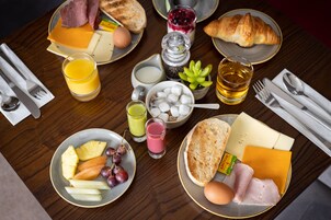 Una tavola imbandita per la colazione con pane tostato, formaggio, prosciutto, uova e frutta. Si vedono anche bicchieri di succo e una brioche.