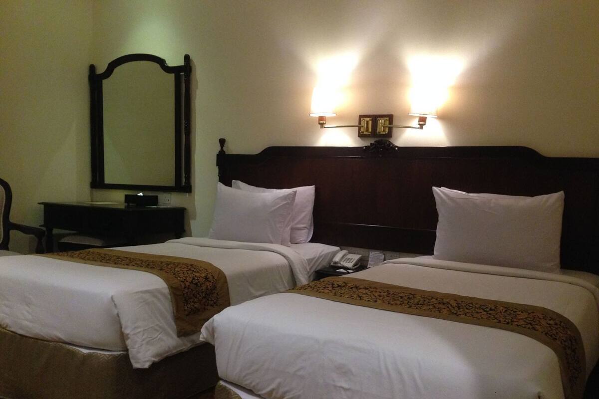 Quarto de hotel com duas camas, uma escrivaninha, uma cadeira, um espelho e uma luminária na parede.