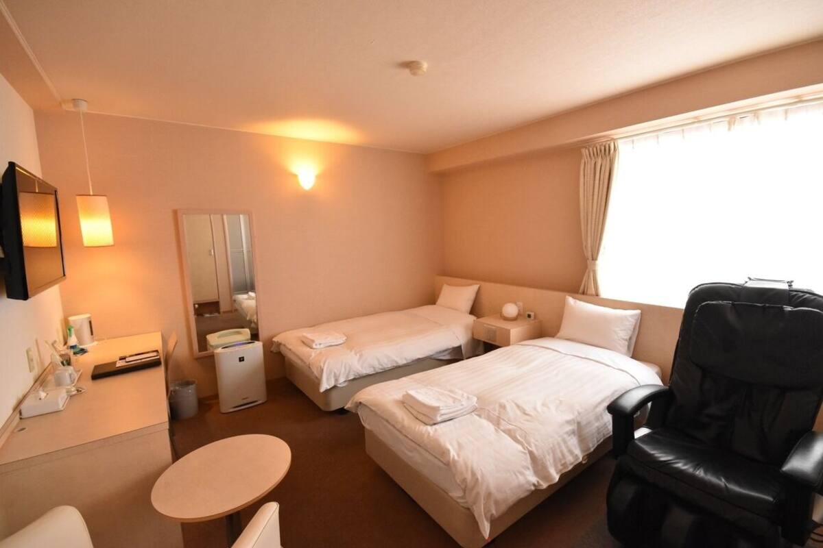 A hotel room with two single beds, a desk with a chair, a round table, a black massage chair, a mirror, and a window with curtains.
