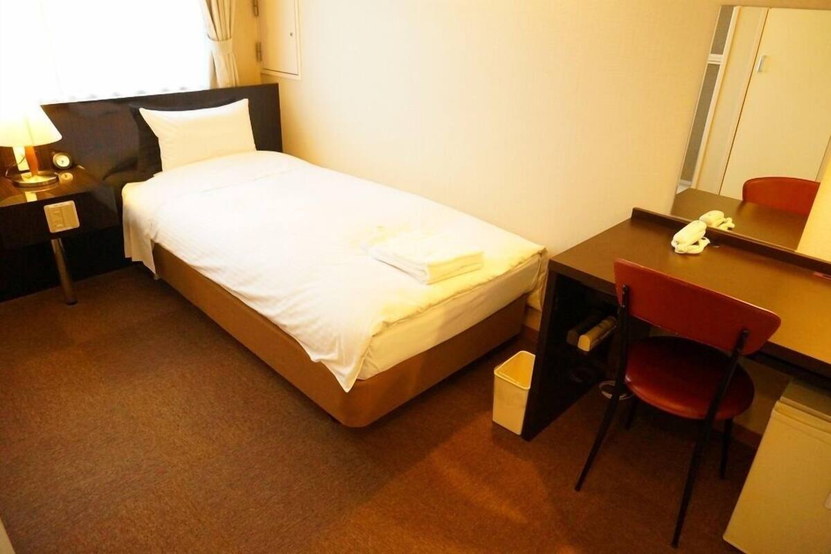 A hotel room with a bed, a desk, a chair, a mirror, a nightstand, and a lamp.