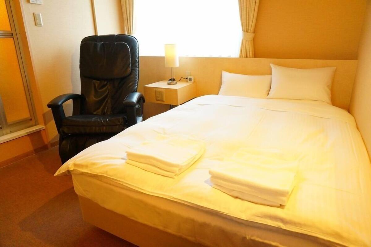 A hotel room with a bed, a chair, a nightstand with a lamp, and a window with curtains.