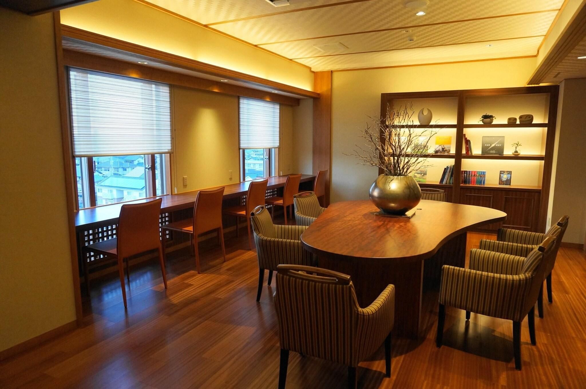 A conference room with a round table, chairs, a bookshelf, and a window with blinds.