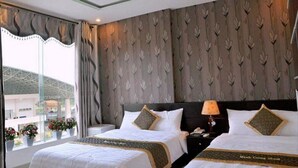 A hotel room with two beds, a nightstand, a lamp, and a window with curtains. - Manh Cuong Hotel (Da Nang)