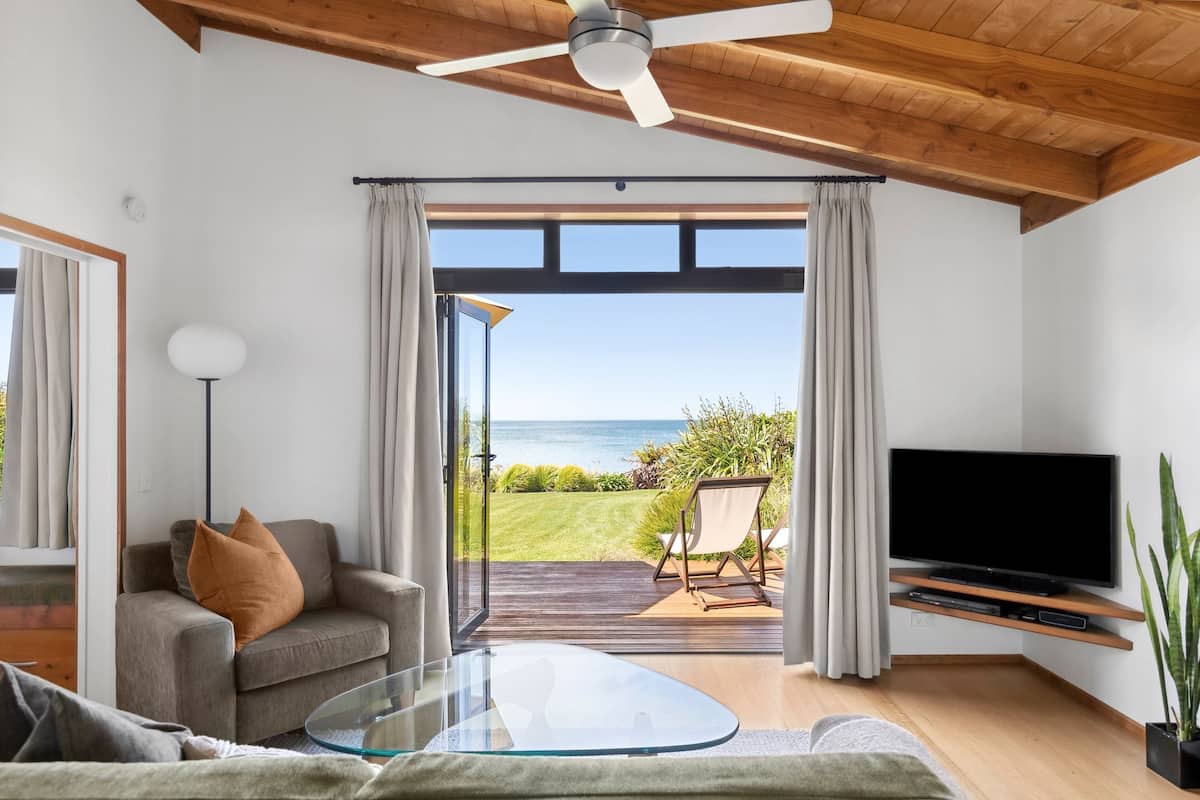 A modern living room with a large TV, a sofa, a glass coffee table, and a view of the ocean through a sliding glass door.