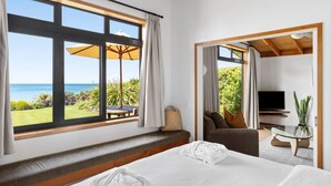 A modern bedroom with a large window overlooking a garden and the sea, a bed, a seating area with a TV, and a small table. - Adrift In Golden Bay (Parapara)