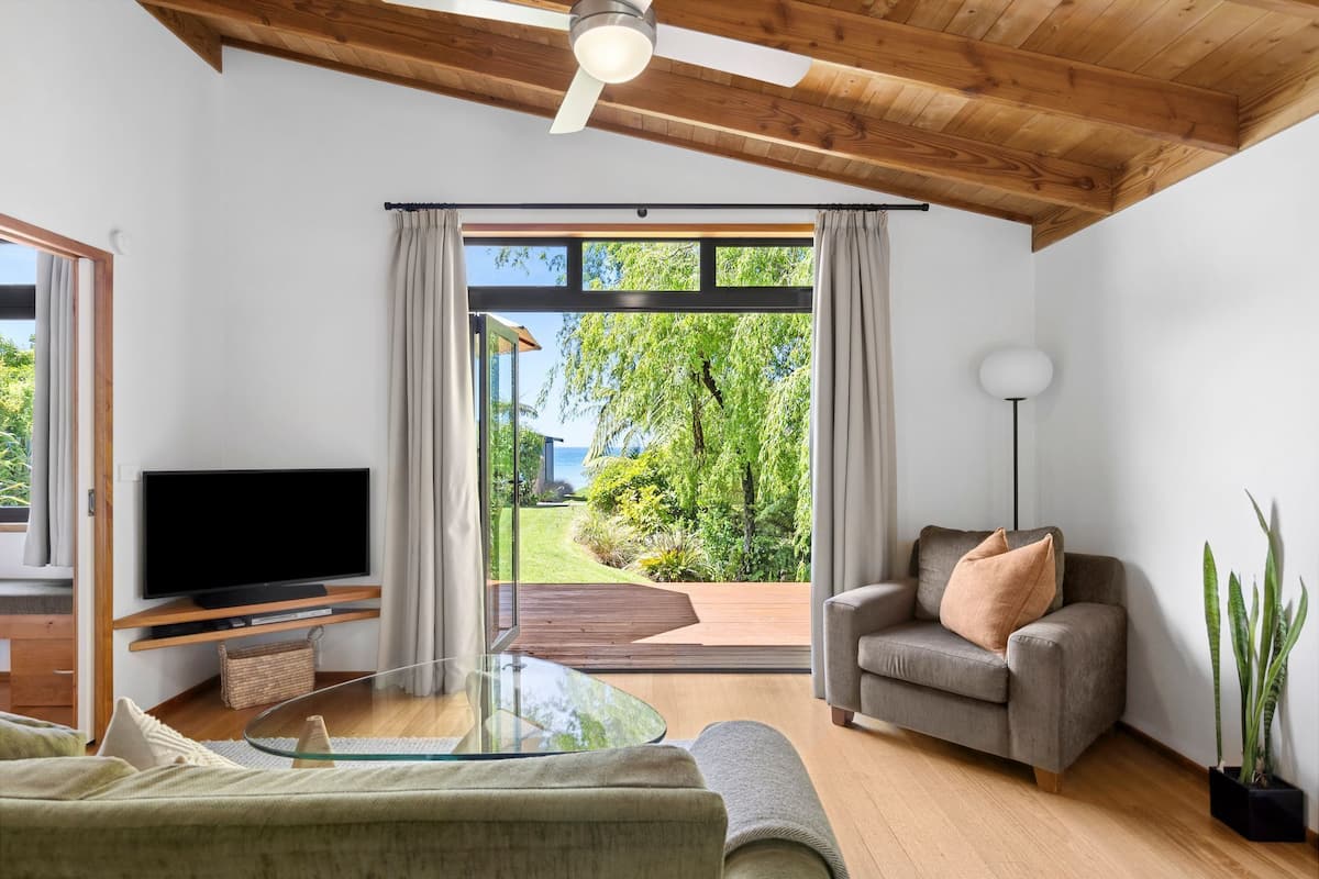 A modern living room with a flat-screen TV, a glass coffee table, a sofa, a grey armchair, and a large sliding glass door leading to a deck with a view of greenery and a building.