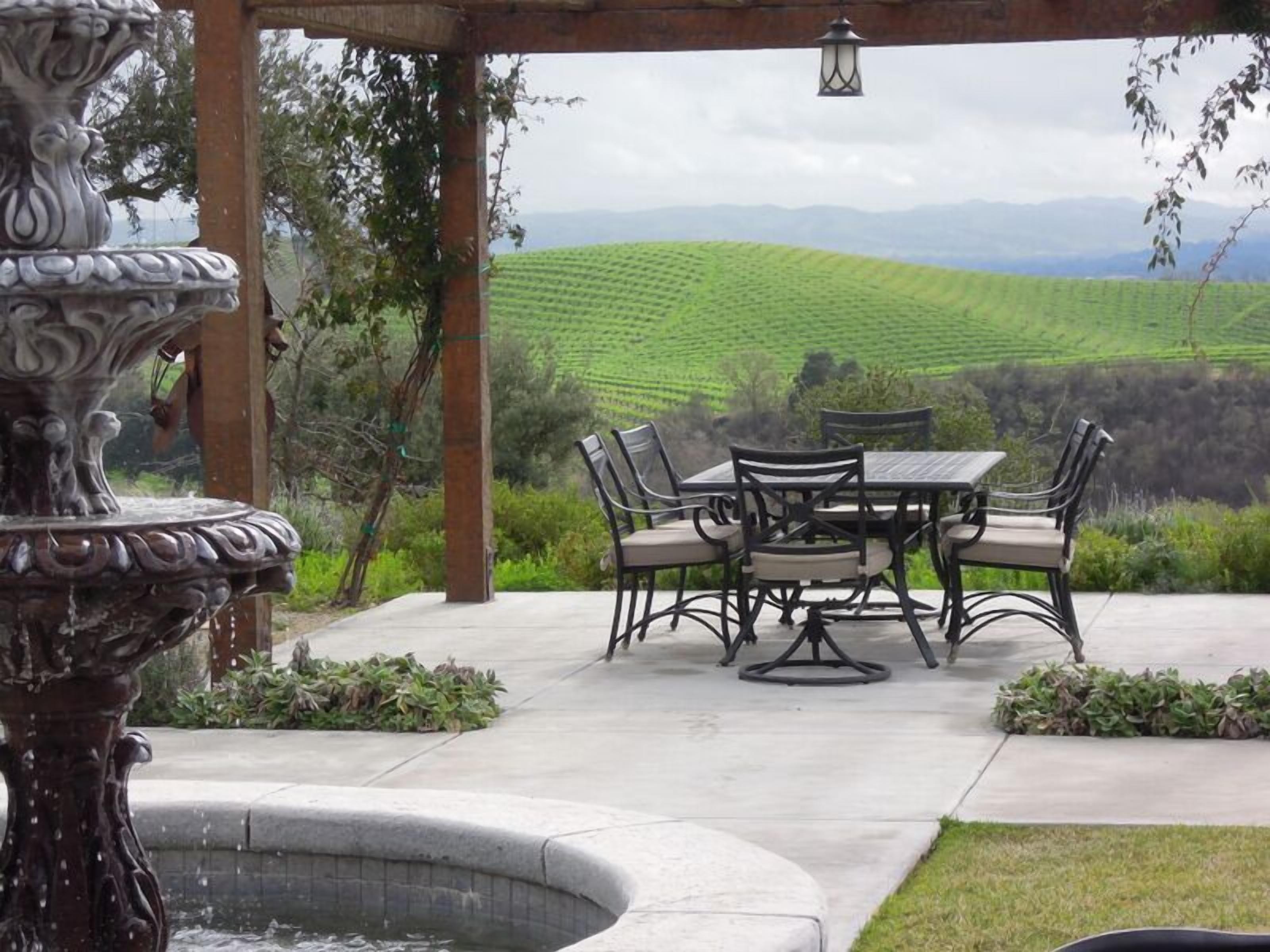 An outdoor patio with a table and chairs, a decorative fountain, and a view of green fields.