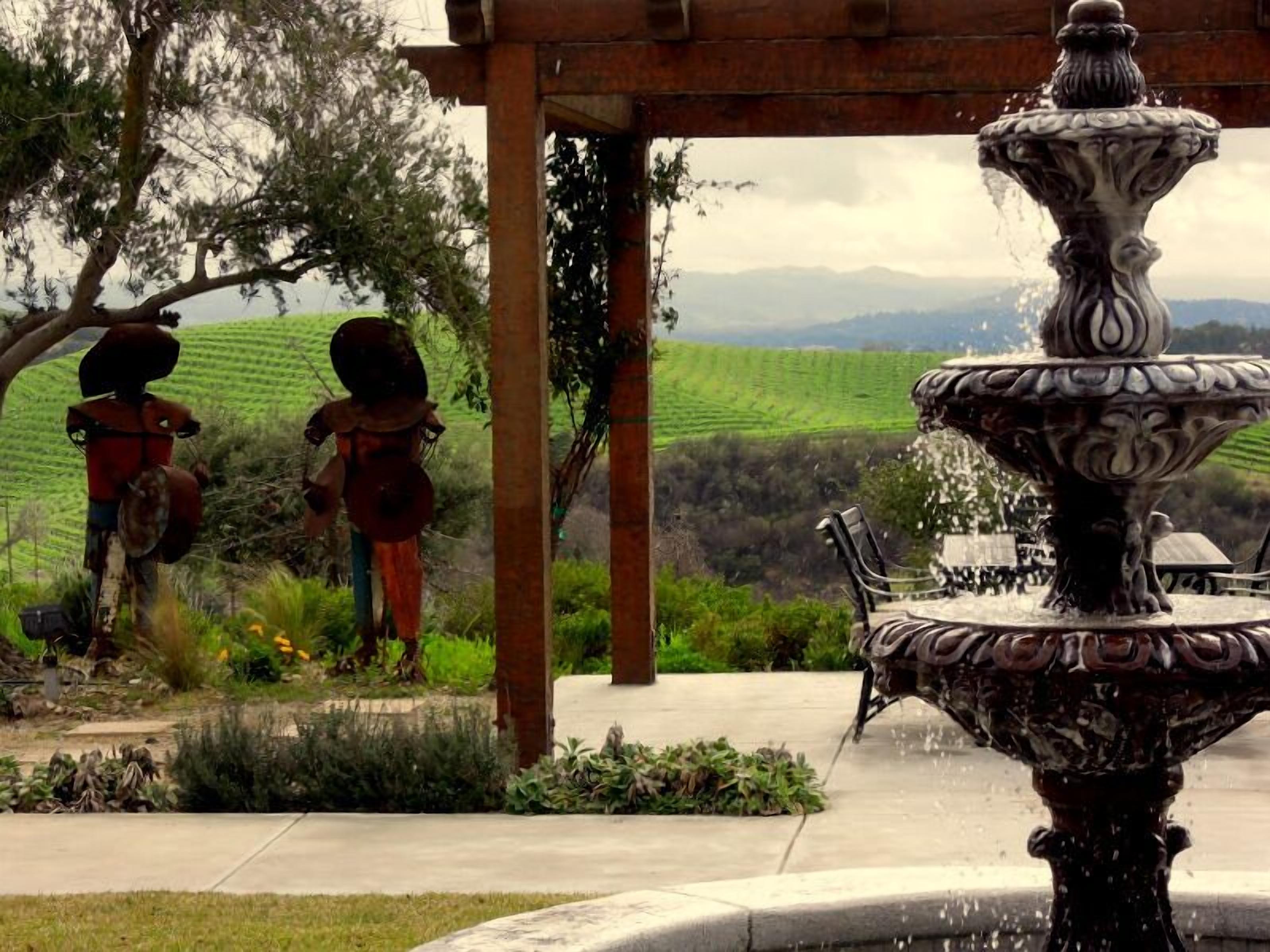 A fountain with intricate design, a pergola structure, and a view of green hills in the background.