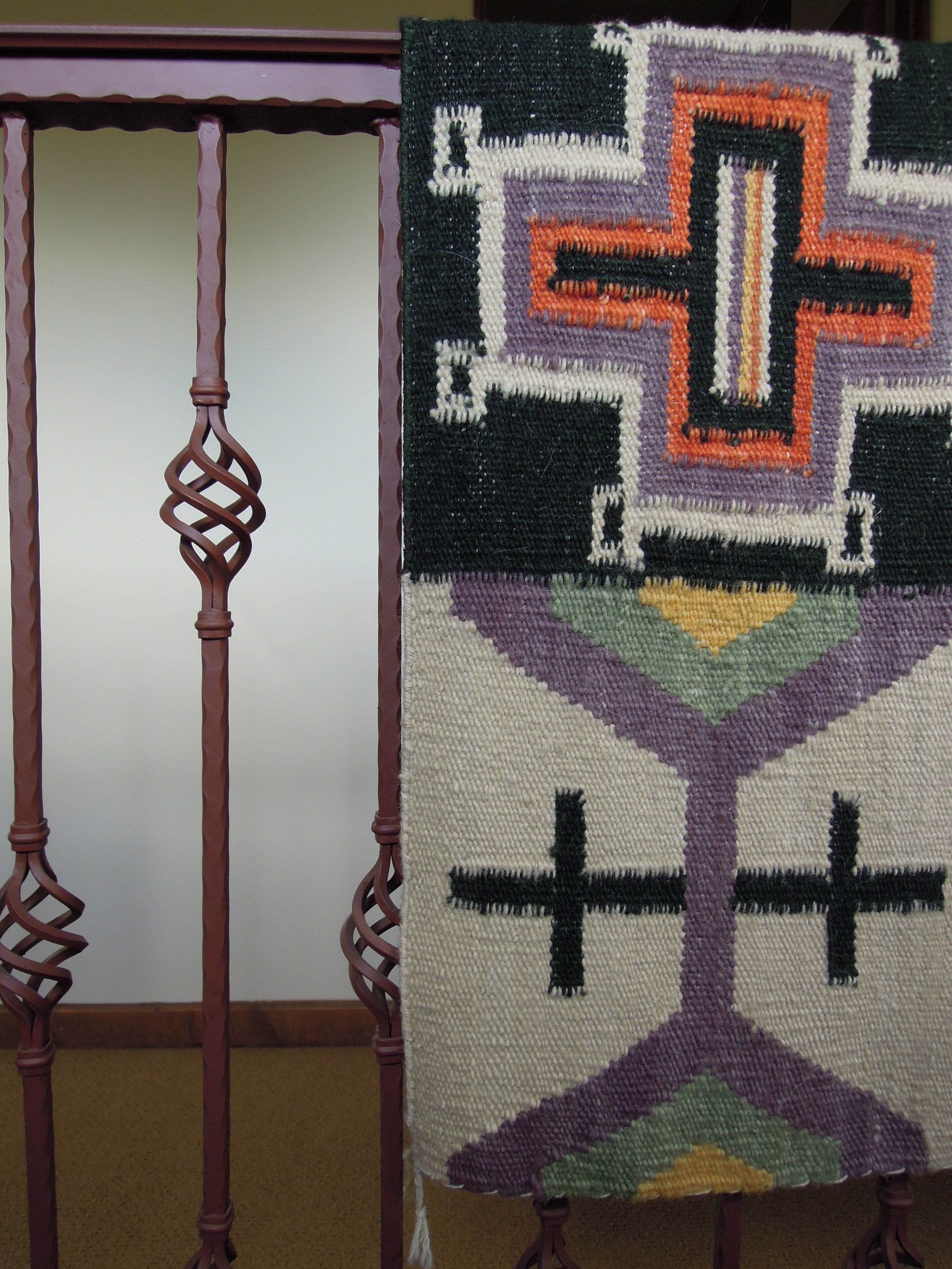 A handwoven textile with geometric patterns and a cross design, displayed on a wooden chair with a simple, elegant backrest.