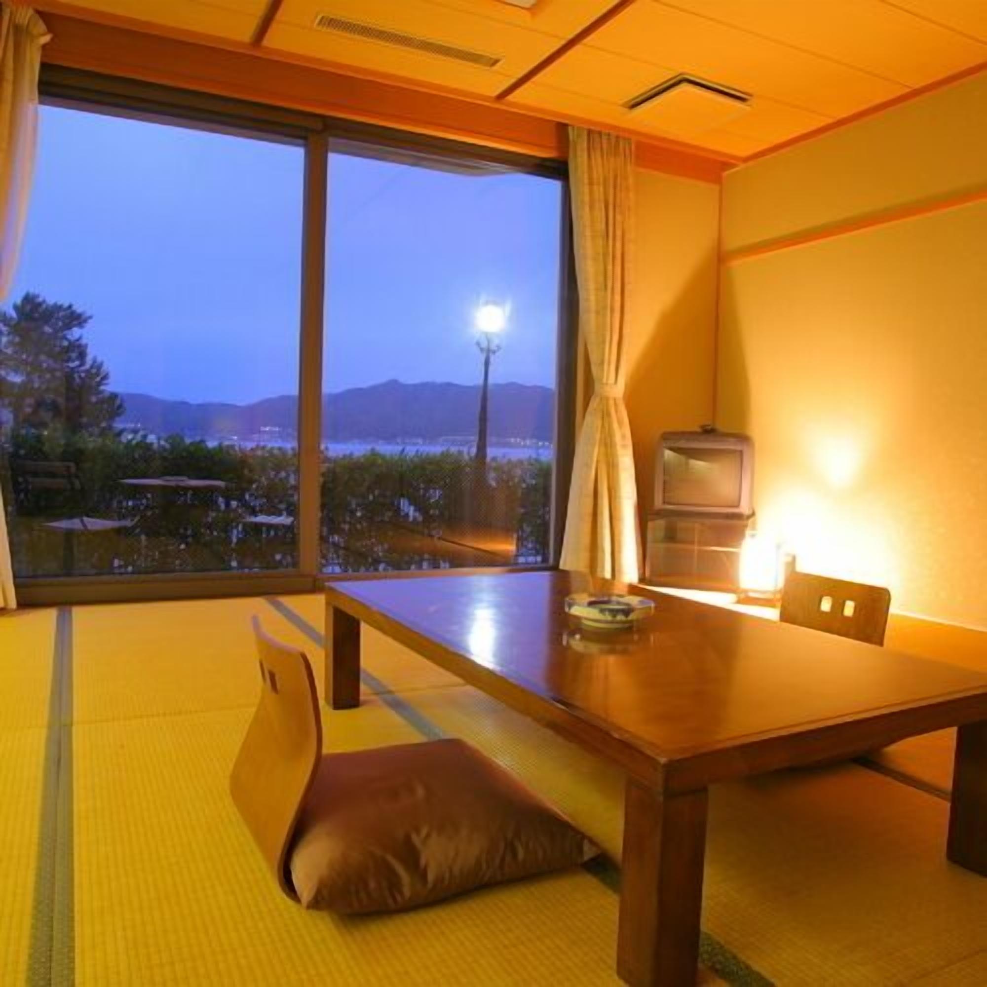 Una habitación japonesa tradicional con suelo de tatami, una mesa baja, cojines, un televisor y vistas a un paisaje urbano al atardecer.