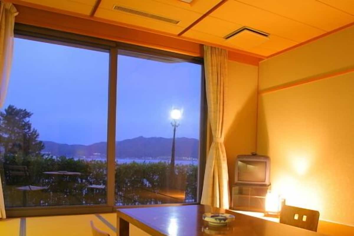 A traditional Japanese room with tatami flooring, a low table, cushions, a television, and a view of a cityscape at dusk.