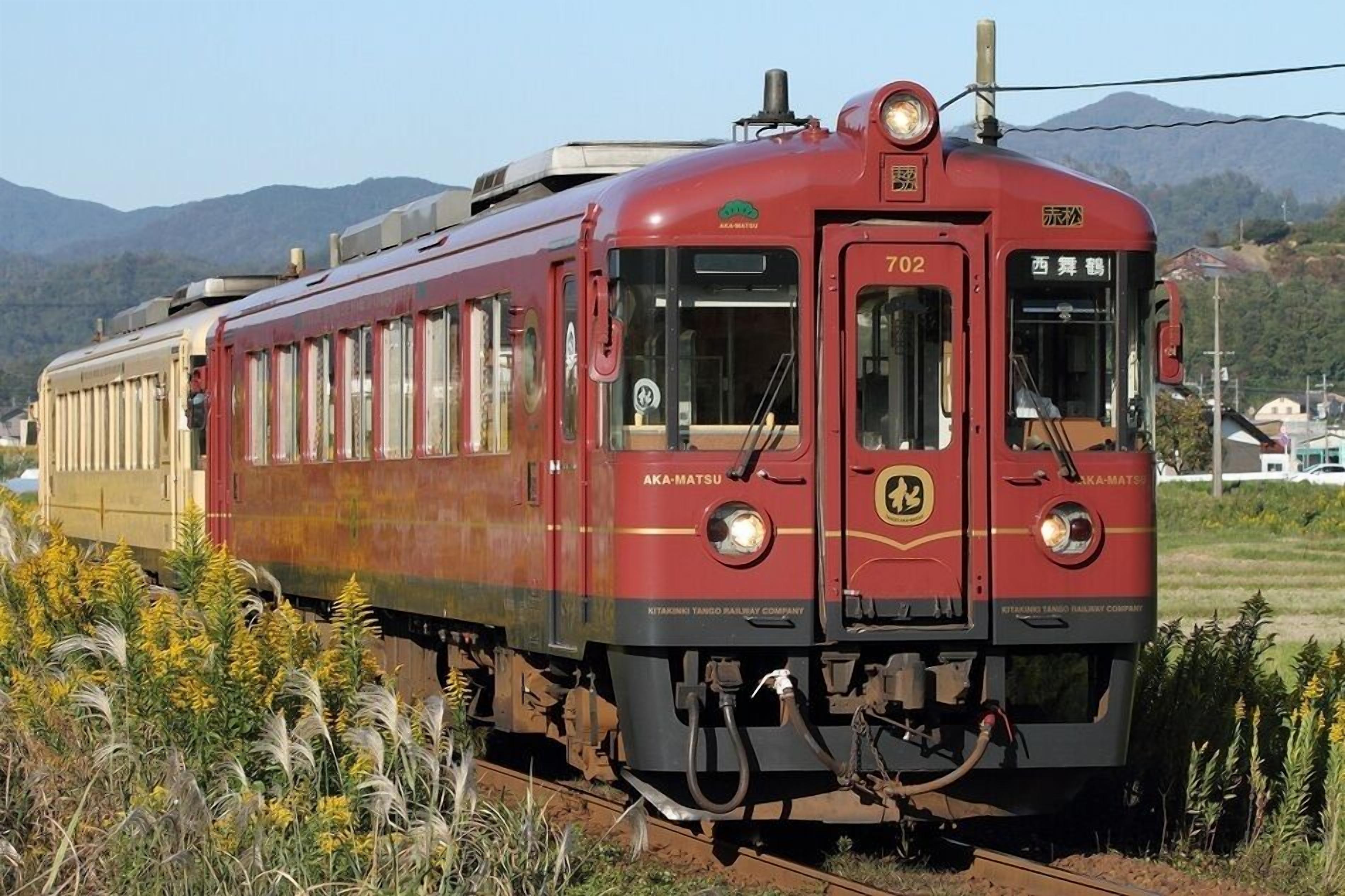 A red and beige train with the number 702, traveling on tracks with mountains in the background.