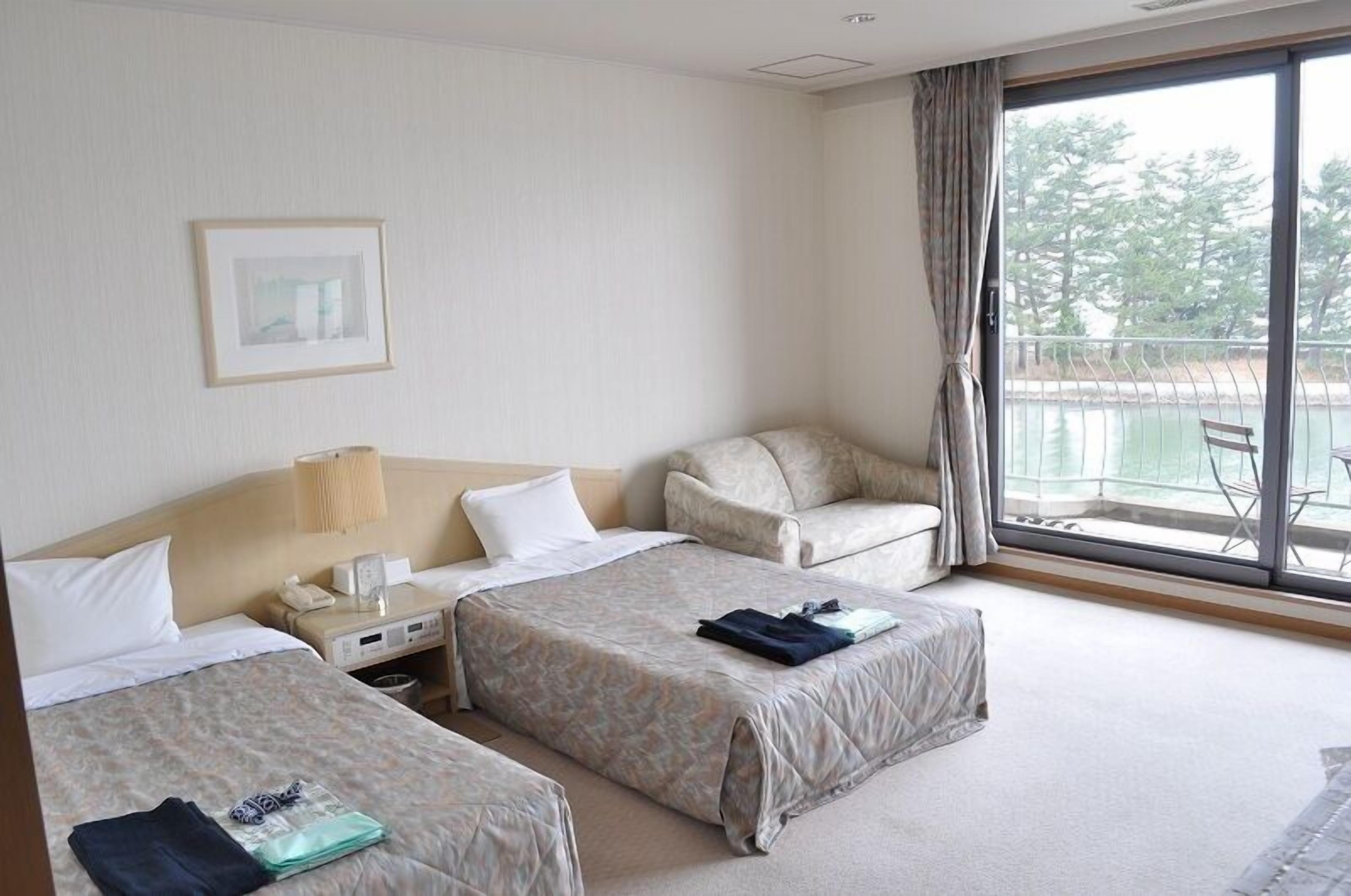 Habitación de hotel con dos camas, un escritorio con lámpara, una silla y un ventanal con vistas a árboles y agua.