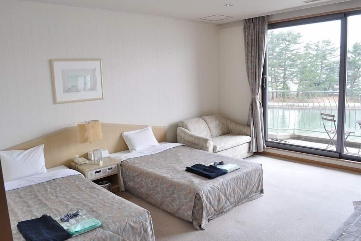 A hotel room with two beds, a desk with a lamp, a chair, and a large window with a view of trees and water.