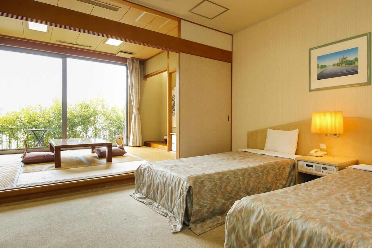 A hotel room with two beds, a desk, a chair, a window with a view, a painting on the wall, and a bedside lamp.