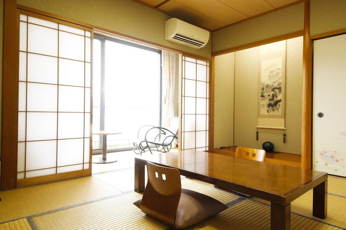 A traditional Japanese room with tatami flooring, a low table, a chair, sliding doors, and a mounted air conditioner.