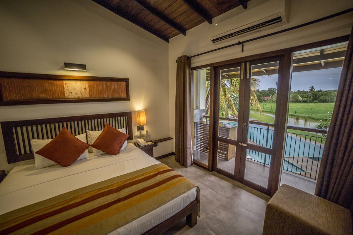 A hotel room with a large bed, a nightstand with a lamp, a wooden headboard, and a view of a pool and greenery through a sliding glass door.