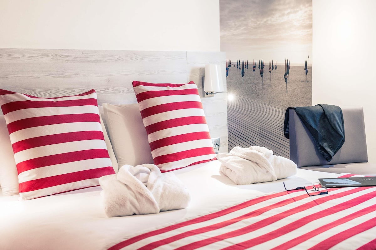 A hotel room with a neatly made bed, striped pillows, a towel, a jacket, and a tablet on the bedside table.