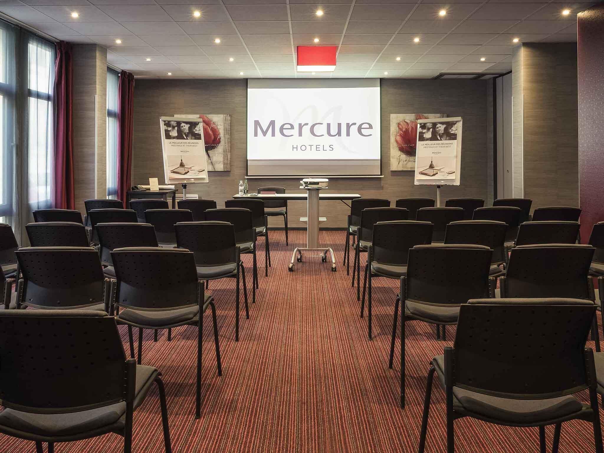 A conference room with rows of chairs, a podium, and a large screen displaying 'Mercure Hotels' branding.