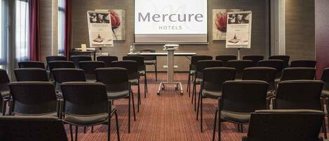 A conference room with rows of chairs, a podium, and a large screen displaying 'Mercure Hotels' branding.