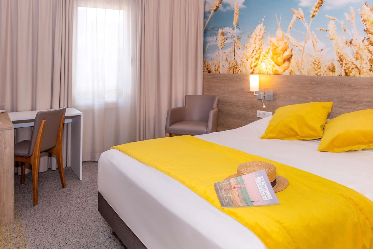 A hotel room with a bed, a desk, a chair, a lamp, and a wall mural.