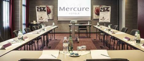 A conference room with a large screen displaying 'Mercure HOTELS' and a presentation. Tables are set with water bottles and notepads.