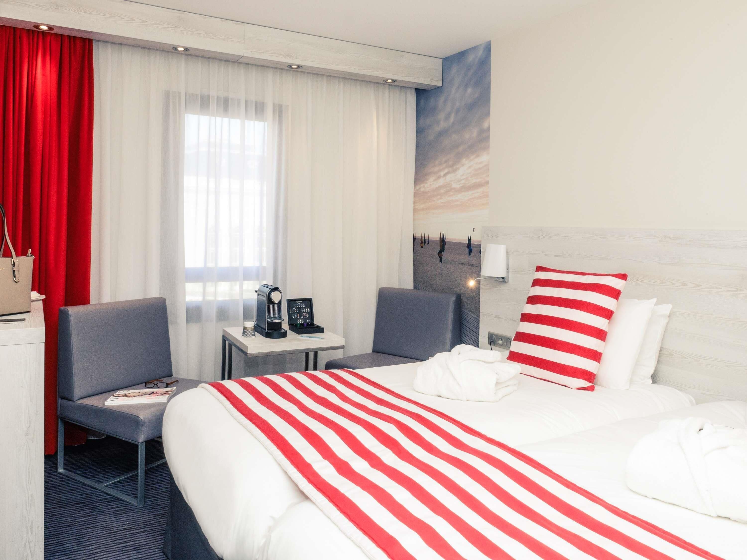 Habitación de hotel con cama, escritorio con cafetera, silla y almohada a rayas roja y blanca.