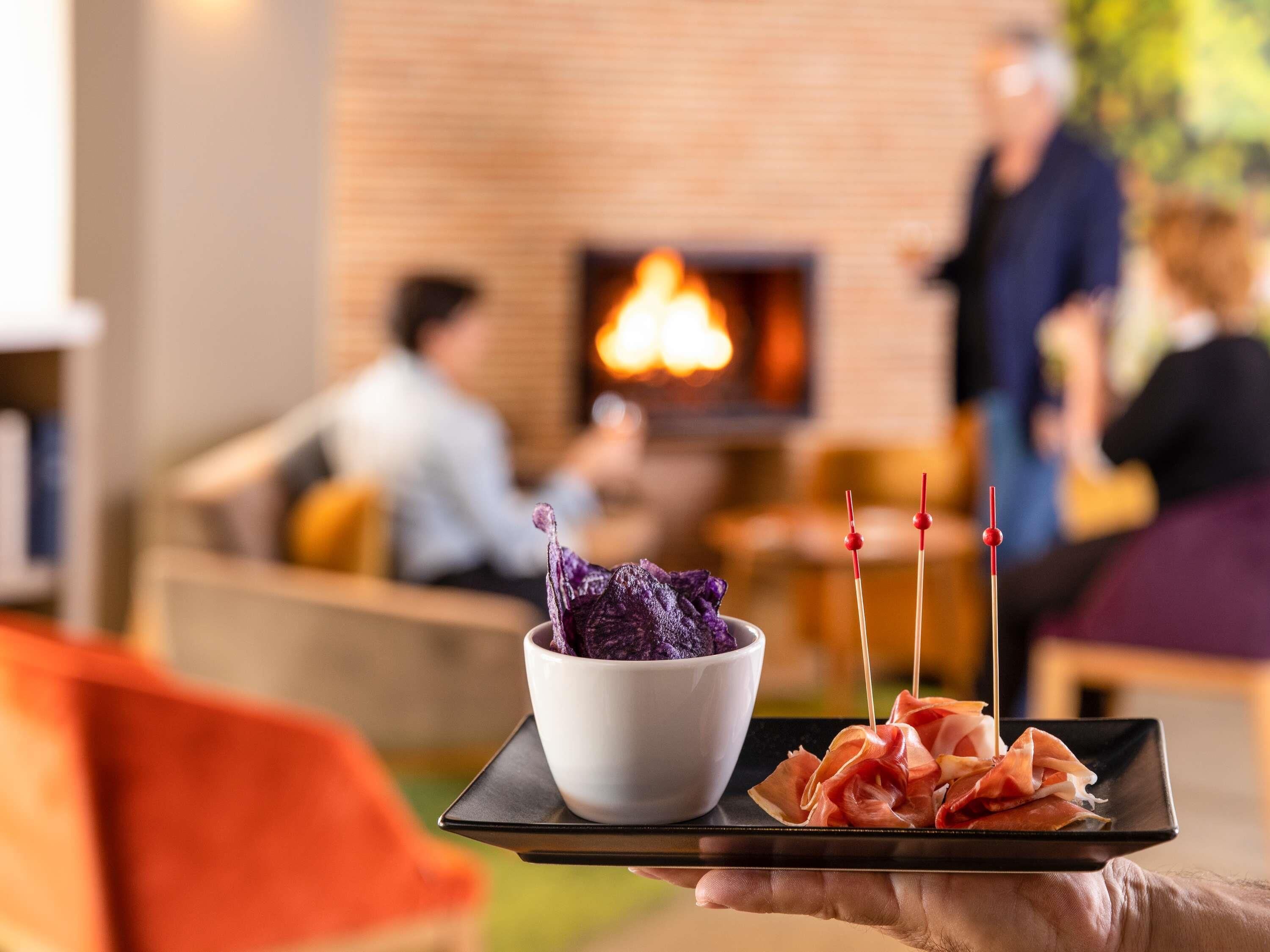 A person holding a tray with a bowl of purple food and sliced meat, with a fireplace in the background.