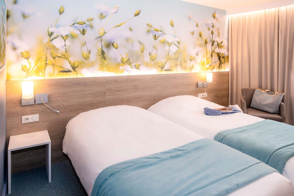 A hotel room with two beds, a nightstand, a lamp, and a wall-mounted light fixture.
