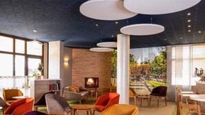 A modern lounge area with a fireplace, comfortable seating, and a large window providing natural light. - Mercure Trouville-sur-mer (Trouville-sur-Mer)