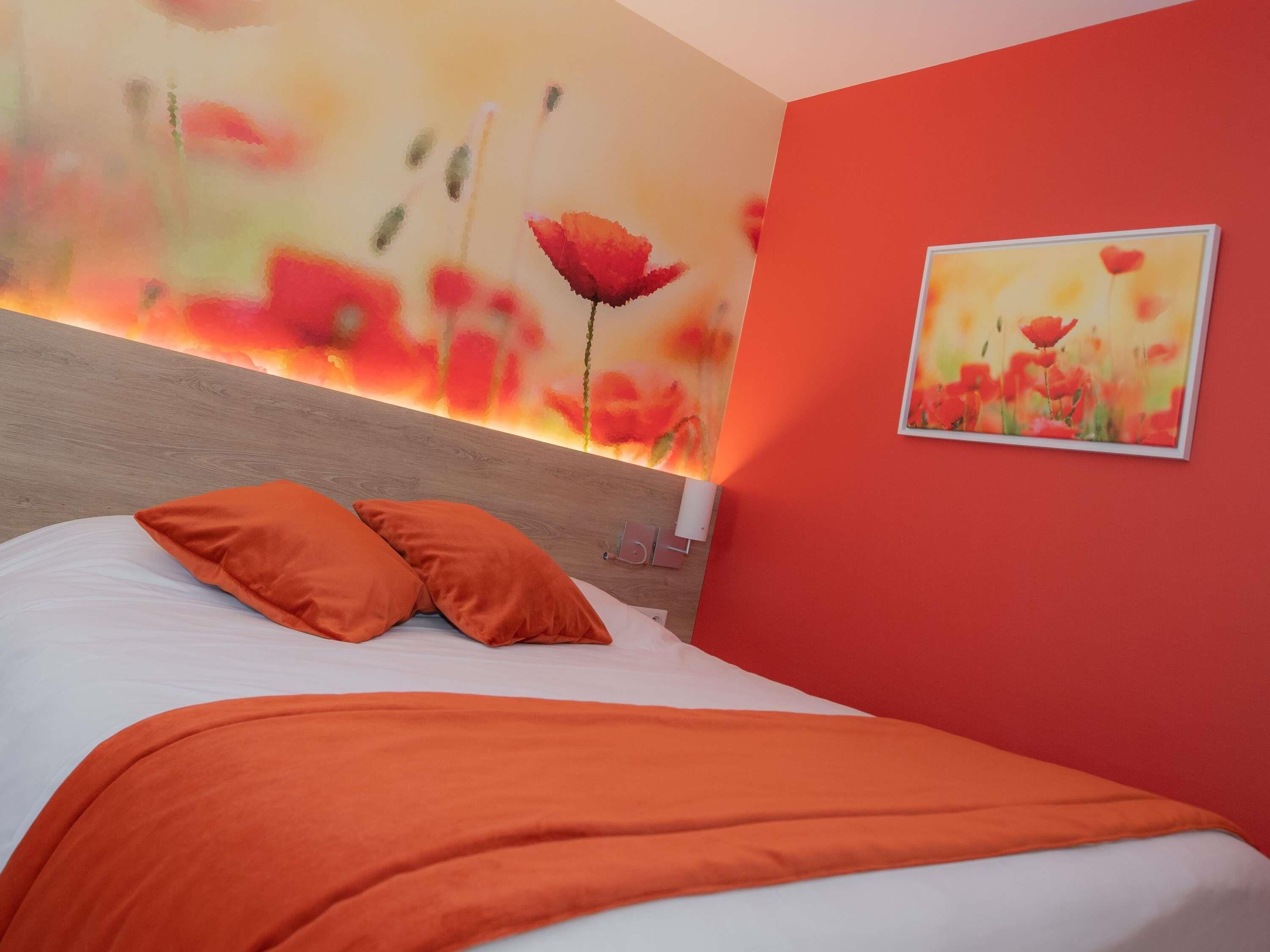 Een hotelkamer met een bed, oranje kussens, een rode muur en een bloemschilderij.