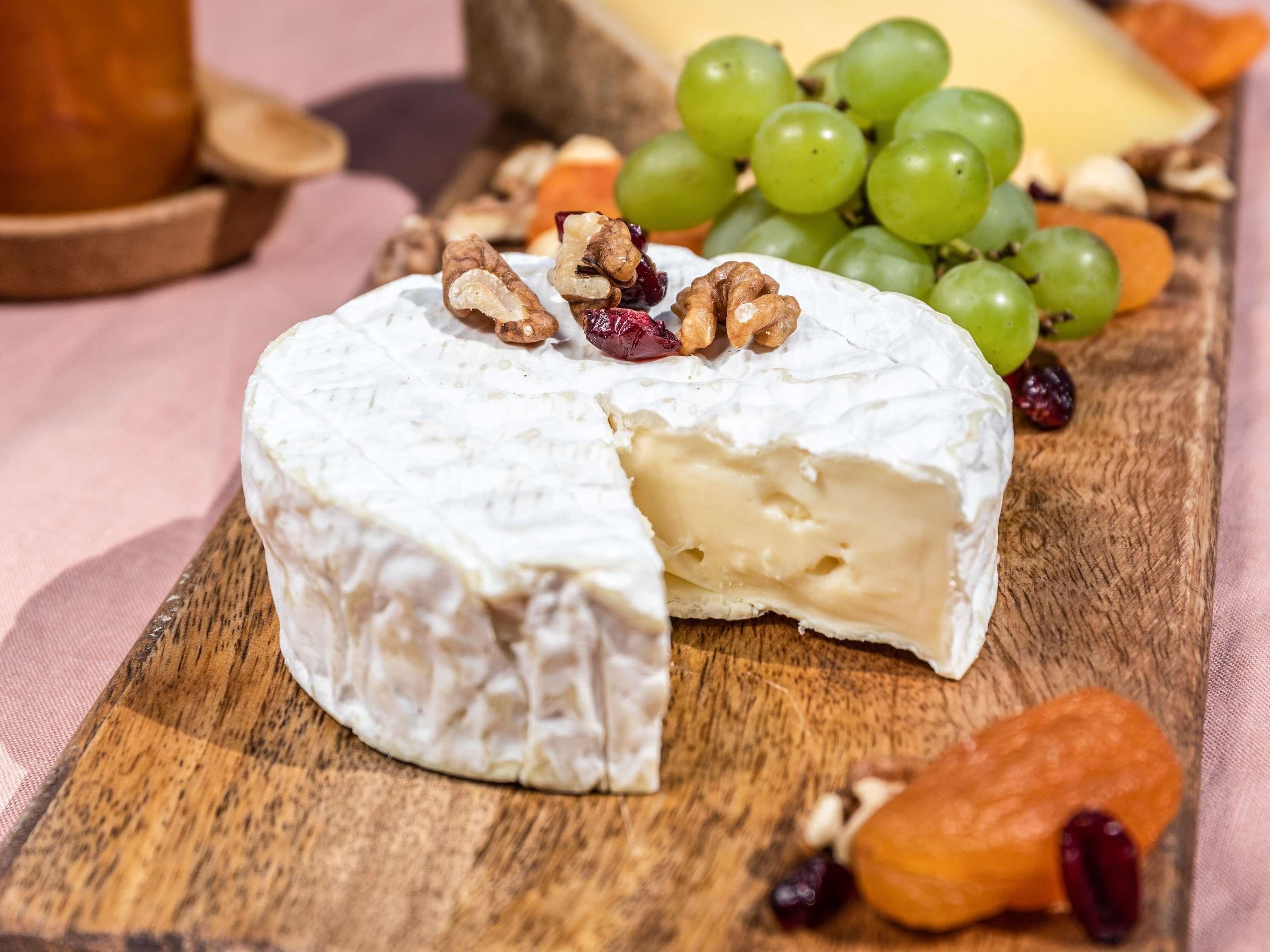 A round cheese wheel with a cut section, surrounded by grapes, nuts, and dried fruits on a wooden board.