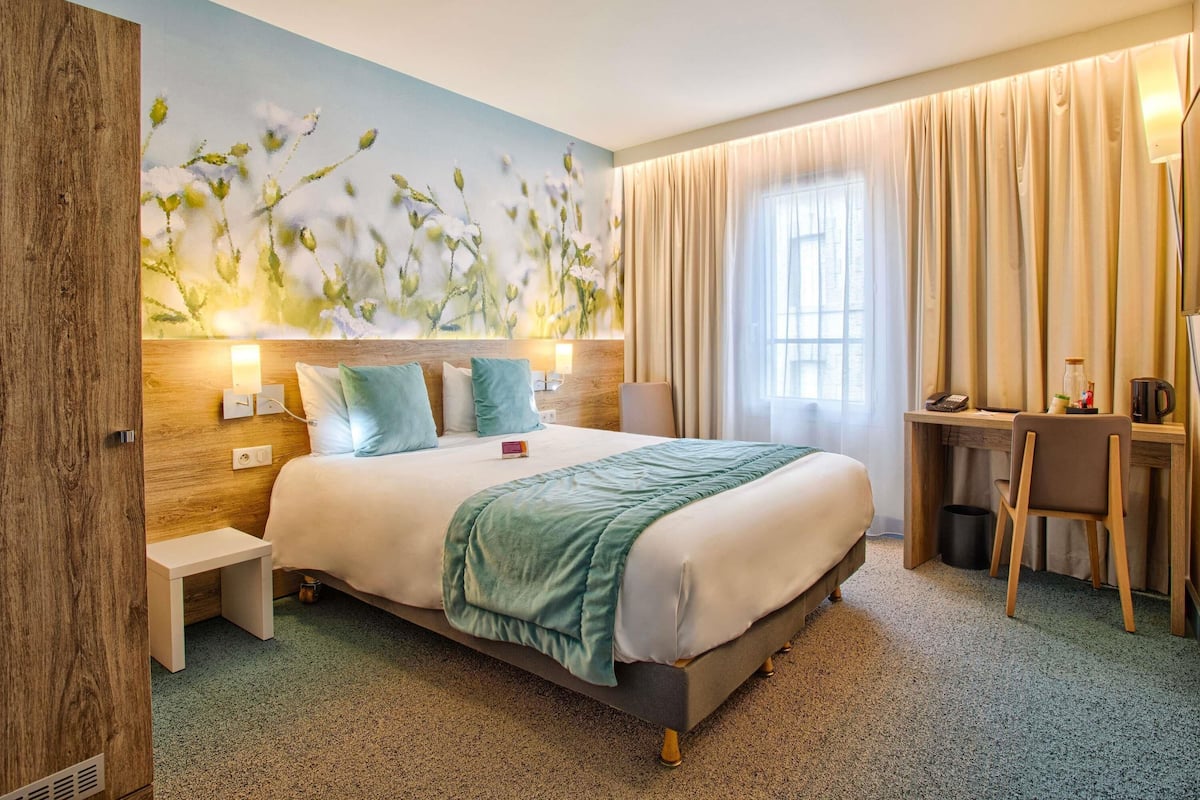 A hotel room with a bed, bedside table, lamp, and a desk with a chair.
