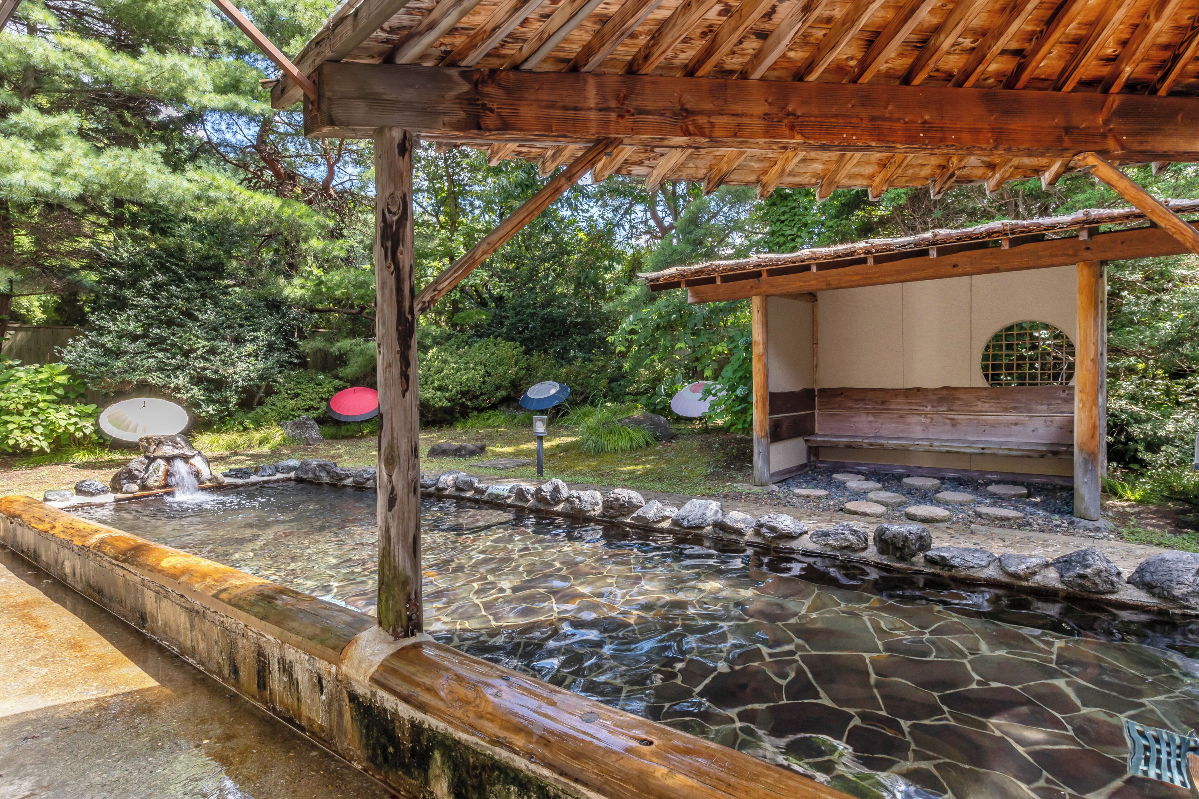 Un onsen japonés tradicional con estructura de madera, cuenca de piedra y vegetación circundante.