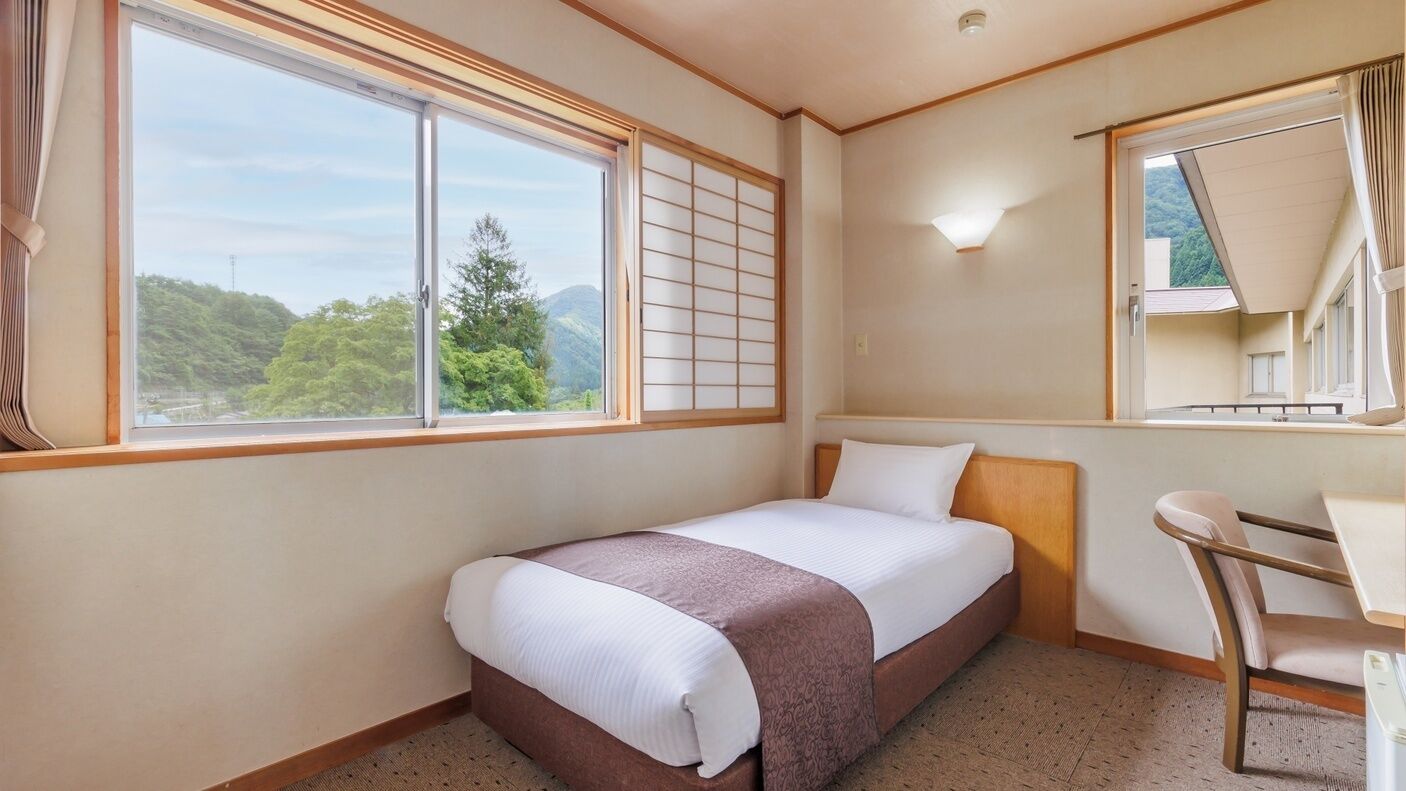 A hotel room with a bed, a desk, a chair, a window with a view of trees, and a mirror.