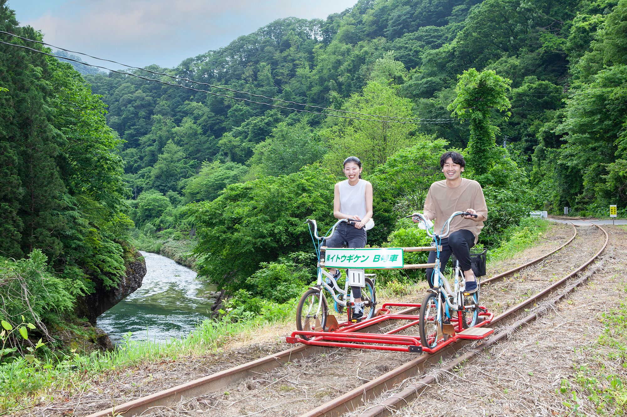 Dos personas montando en bicicleta sobre vías de tren, rodeadas de abundante vegetación y un río.