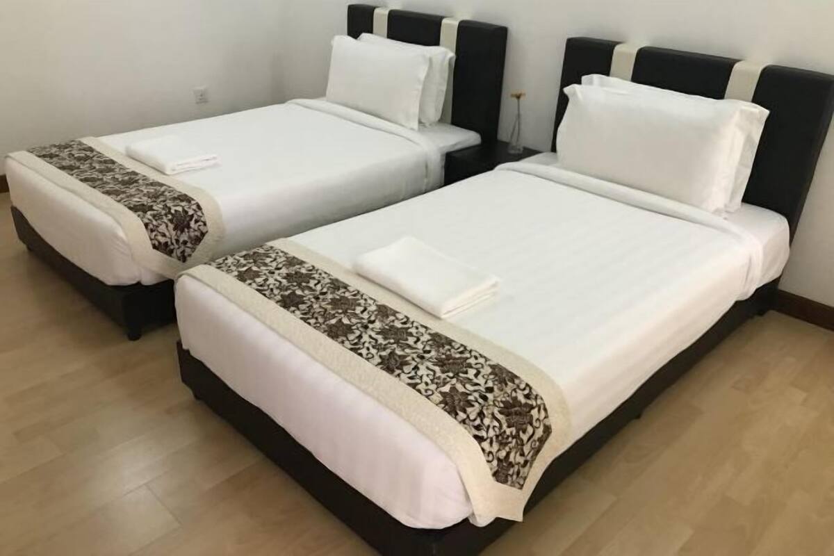 Dos camas individuales con cabeceros negros y ropa de cama blanca, un suelo de madera y una mesita de noche pequeña con una lámpara.