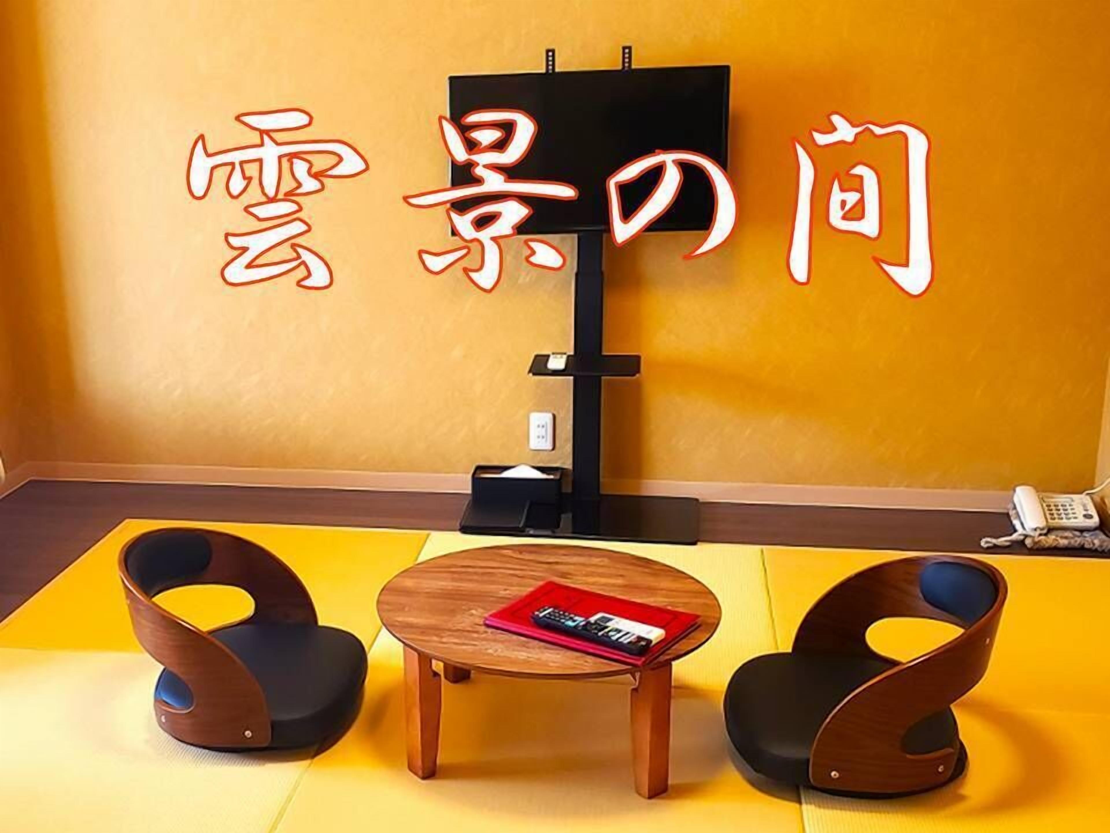 Une pièce avec un sol jaune, une table en bois sur laquelle se trouve un magazine, deux chaises, un meuble pour la télévision et un téléphone fixé au mur.