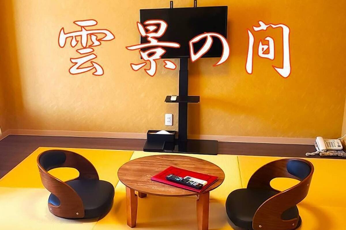 A room with a yellow floor, a wooden table with a magazine, two chairs, a TV stand, and a phone on the wall.