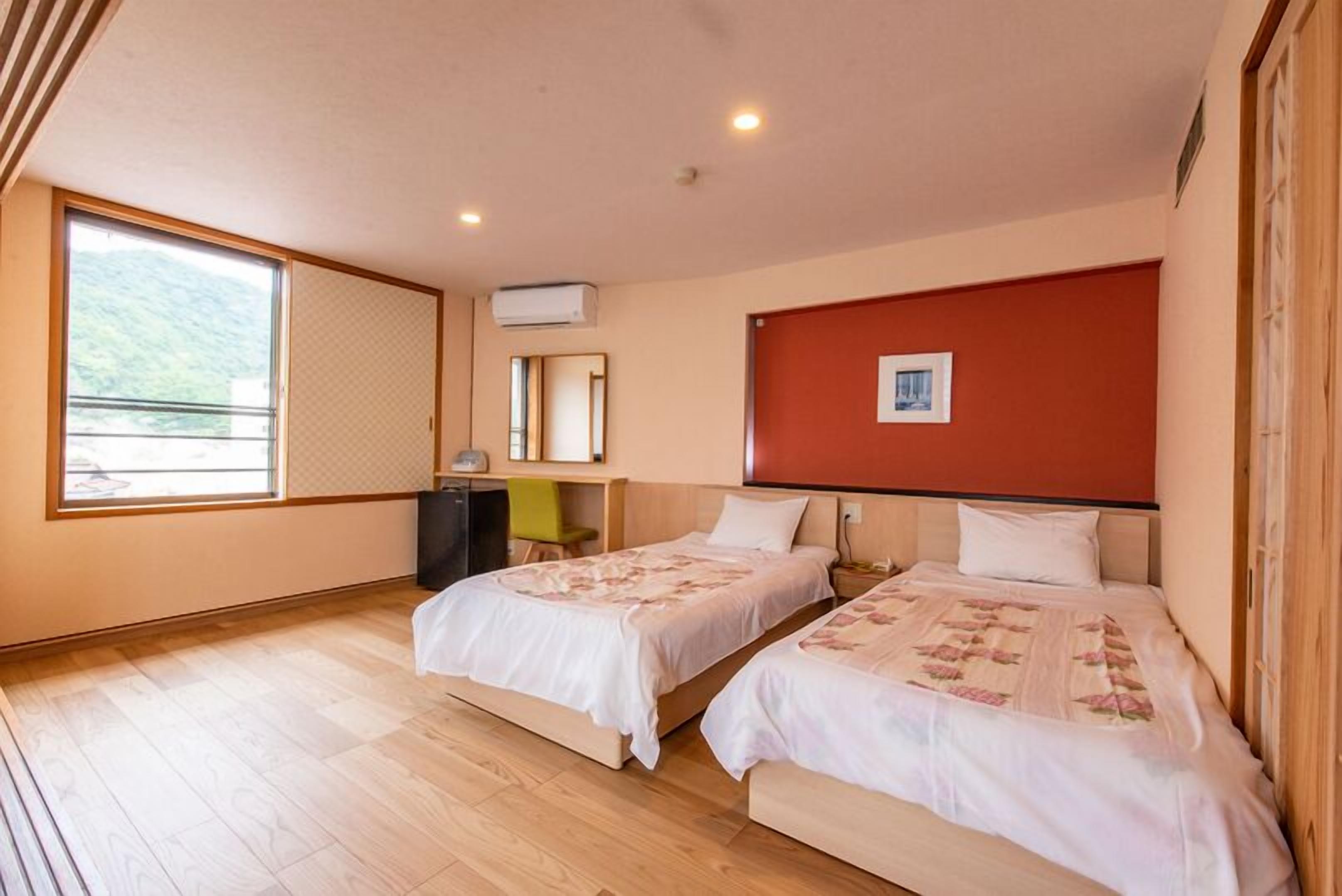 Une chambre d’hôtel avec deux lits, un bureau, une chaise, une fenêtre donnant sur un beau paysage et un mur d’accent rouge.