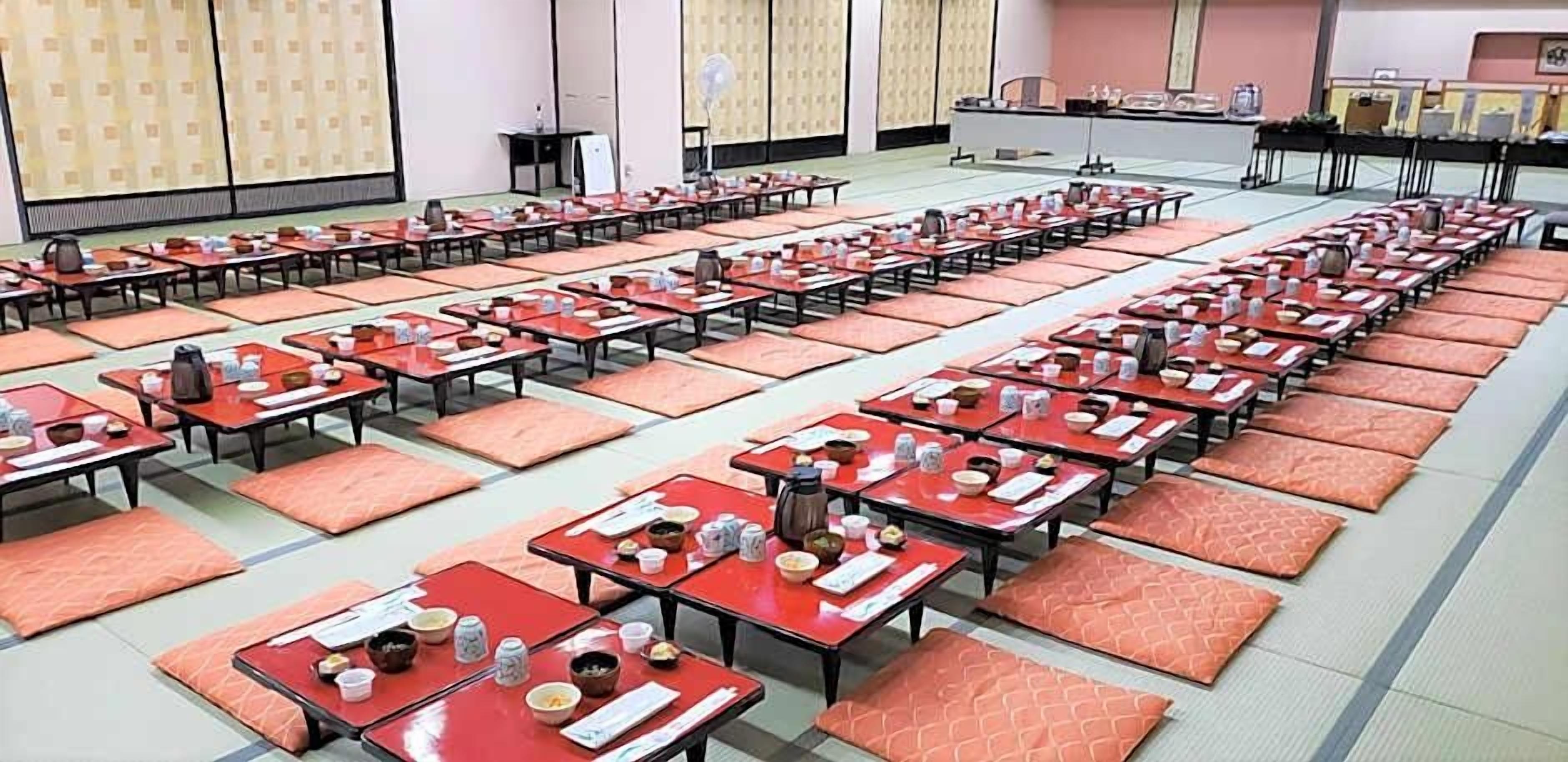 Un salón amplio con filas de mesas y sillas, algunas con objetos encima, y un escenario al fondo.
