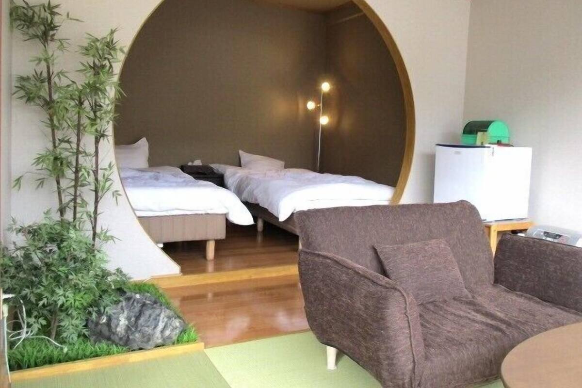 A room with two beds, a sofa, a small table, a refrigerator, and a wall-mounted light fixture.