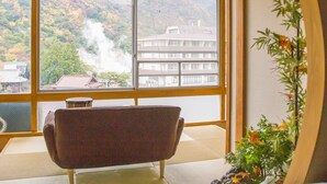 A room with tatami flooring, a traditional Japanese chair, a large window with a view of a mountain and a building, and a bamboo plant. - Unzen Sky (UNZEN)