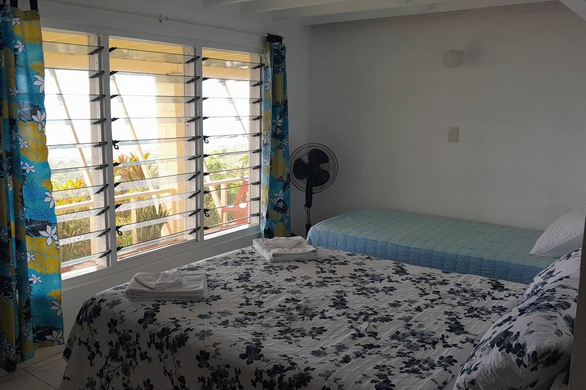 A bedroom with a bed, a standing fan, and a window with curtains.
