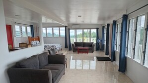 A spacious living area with a sofa, a small table, and a bed with a floral bedspread. There are large windows with curtains, a fan, and a view of greenery outside. - Dave Parker's Eco Lodge (Apia)