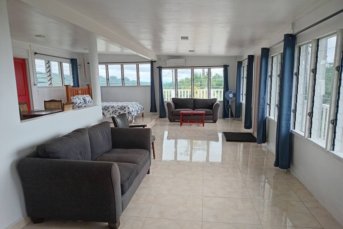 A spacious living area with a sofa, a small table, and a bed with a floral bedspread. There are large windows with curtains, a fan, and a view of greenery outside.