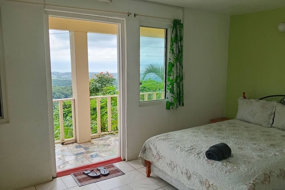 A bedroom with a bed, a window with a view of greenery, and a balcony.