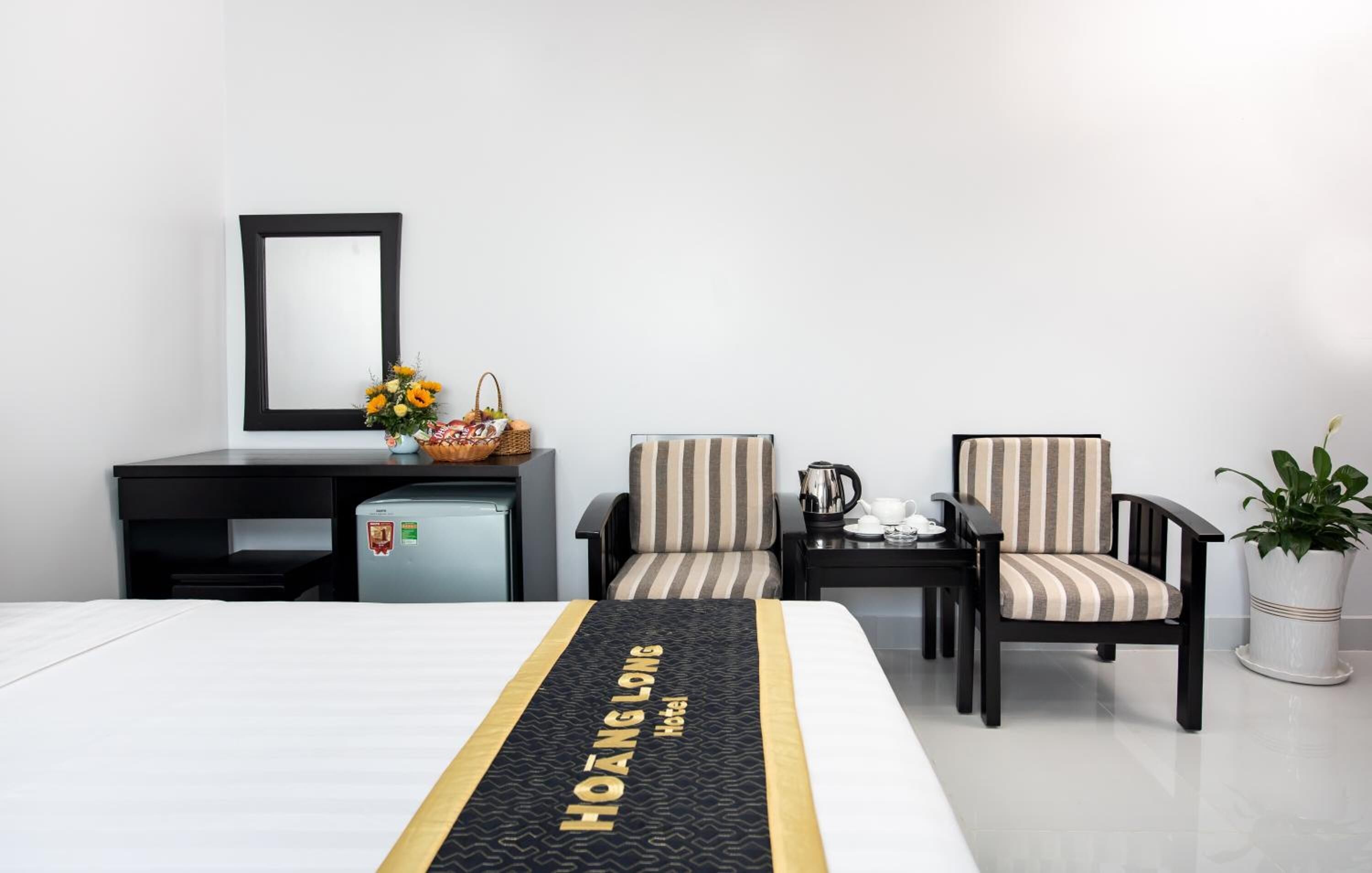 Une chambre d’hôtel avec un lit, deux chaises à rayures, une petite table avec une bouilloire, une plante et un réfrigérateur.