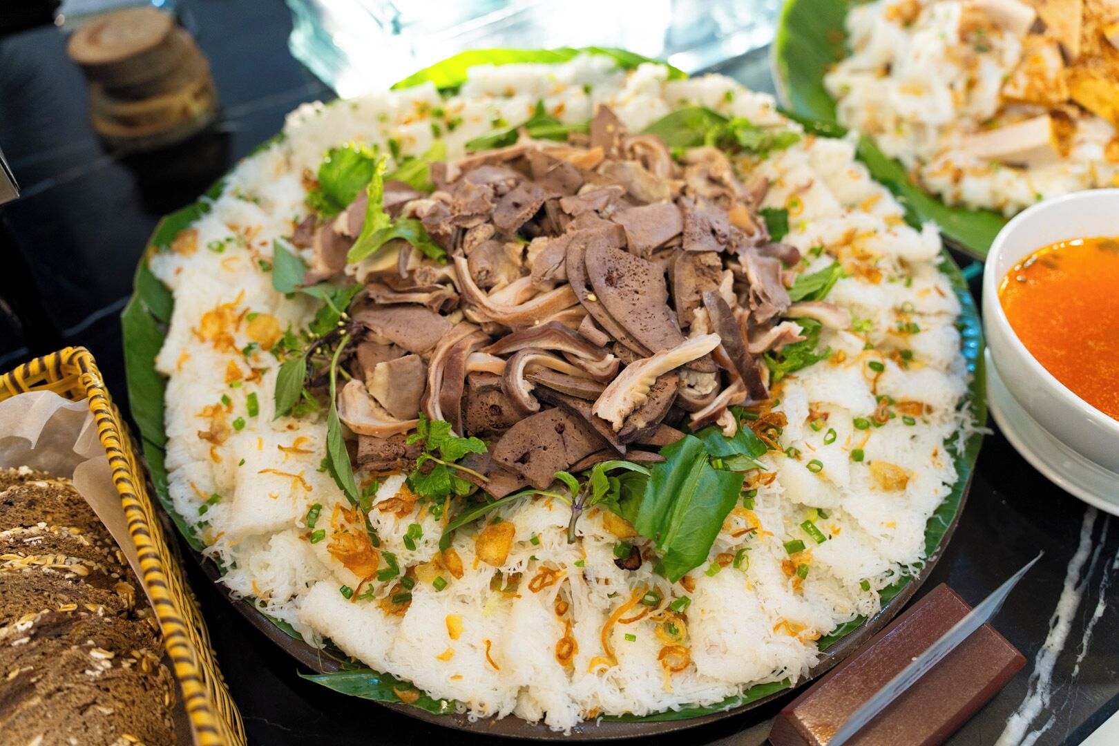 A plate of white rice topped with beef slices and garnished with green onions and herbs, accompanied by a bowl of sauce.
