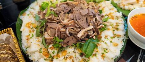 A plate of white rice topped with beef slices and garnished with green onions and herbs, accompanied by a bowl of sauce.
